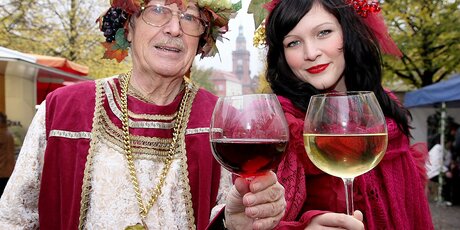 Eine frau und ein Mann in Trachten stoßen mit einem Glas Rot- und Weißwein an.