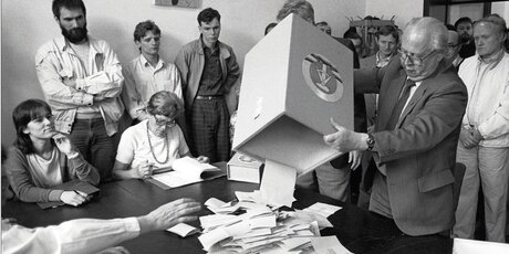 Schwarz-Weiß-Foto. Wahlhelfer beim ausleeren einer Wahlurne unter Beobachtung von Bürgern.